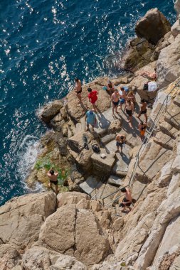 Dubrovnik. Hırvatistan - 18 Kasım 2025: Hırvatistan 'ın Dalmaçya kentindeki bir uçurum plajında yaz tatili sahnesi. Tanınmayan insanlar köpüklü mavi suyun yanındaki kayalıklarda dinleniyorlar. Seyahat ve eğlence için ideal..