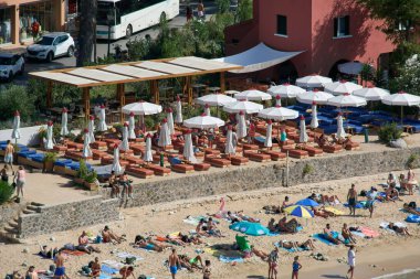 Korfu. Yunanistan - 15 Aralık 2025: Çok sayıda turistin kum ve çakıl üzerinde güneşlendiği organize kıyı sahnesi toplu turizm ve tatil tatillerini ön plana çıkarıyor.