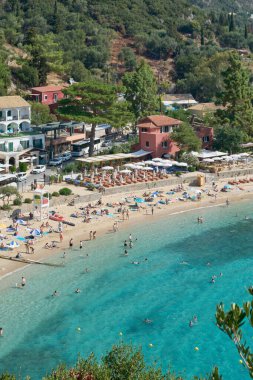 Korfu. Yunanistan - 15 Aralık 2025: Canlı yaz sahnesi, turistlerin yüzdüğü ve güneşlendiği yoğun bir körfezi çevreleyen terasotta renkli binaların arkasındaki yeşil tepeler.