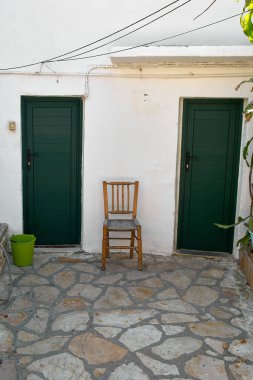 Akdeniz 'deki bir köyün düzensiz taş zemininde iki koyu yeşil kapısı ve kırsal ahşap bir sandalyesi olan beyaz bir duvarın simetrik yapısı.