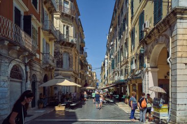 Korfu. Yunanistan - 27 Ocak 2026: Tarihi cephelerin arasına gizlenmiş restoranların masalarında öğle yemeği yiyen eski bir kasaba caddesinin perspektifli manzaralı kent sahnesi.