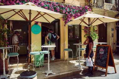 Korfu. Yunanistan - 27 Ocak 2026: Turistler, parlak bir yaz gününde büyük güneş şemsiyeleri altında canlı pembe bougainvillea çiçekleriyle süslenmiş büyüleyici bir zanaatkâr dondurmacının önünde toplandılar.