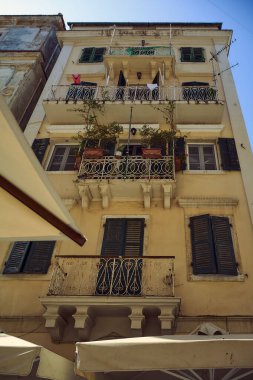 Çok katlı Akdeniz binası ön cephesinde dövülmüş demir parmaklıklar, yeşil kepenkler ve temiz bir gökyüzünün altında dekoratif saksı bitkileri olan geleneksel balkonlar yer alıyor.