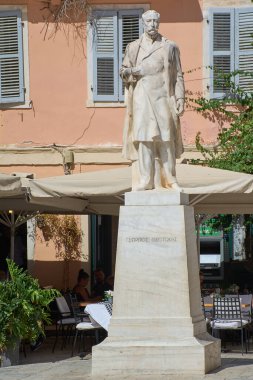 Korfu. Yunanistan - 27 Ocak 2026: Georgios Theotokis 'e adanmış beyaz mermer heykelin dikey çekimi, güneşli bir kasaba meydanında yerel kafe teraslarıyla çevrili duruyor