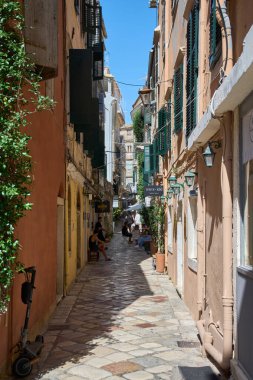 Korfu. Yunanistan - 29 Ocak 2026: İnsanlar butik ve kafelerle dolu dar bir eski kasaba caddesinde oturup yürüyor, rahat turizm, tarihi mimari ve günlük Akdeniz şehir yaşamını yakalıyorlar.