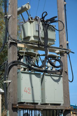 Elektrik güç transformatörü ve kontrol dolapları ahşap elektrik direğine bağlı, açık mavi gökyüzüne karşı ağır kablolar, enerji dağılımını, şebeke altyapısını ve güvenilir kentsel elektrik kaynaklarını sembolize ediyor..