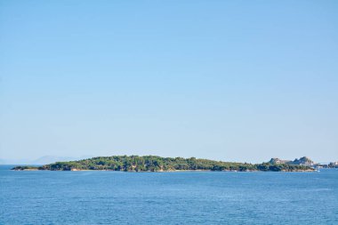 Yeşil bir adanın panoramik deniz manzarası kıyı şeridi boyunca tarihi kaleler, sakin mavi sular ve Akdeniz seyahatini, mirası ve kıyı huzurunu ön plana çıkaran berrak bir gökyüzü.