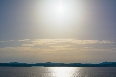 Işıl ışıl deniz yüzeyi yoğun güneş ışığını yansıtıyor berrak bir gökyüzünün altında kıyı siluetleriyle..
