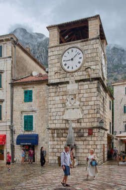 Kotor. Karadağ - 06 Şubat 2026: Eski kentin ana meydanında turistler ve dağlarla birlikte tarihi taş saat kulesi.