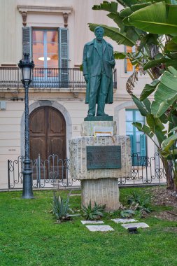 Siteler. İspanya - 19 Şubat 2026: Santiago Rusinol 'un peyzaj bahçesindeki bronz heykeli, tarihi cephe ve palmiye yaprakları kıyı kenti merkezindeki kültürel yapıyı çerçevelemektedir..