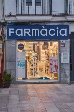 Siteler. İspanya - 22 Şubat 2026: Şehir merkezindeki ticari caddede görünür tabela ve sağlık ürünleri vitrinleriyle aydınlatılmış Farmacia mağazası