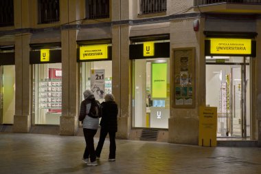 Sitges, İspanya - 02 Mart 2026: Yayalar, geceleri parlak sarı ticari işaretler ve aydınlatılmış pencere vitrinleriyle modern bir Optica y Audiologia Universitaria mağazasının önünden geçiyor.