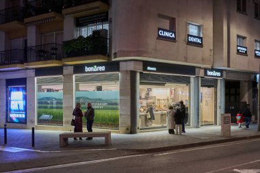 Siteler. İspanya - 12 Mart 2026: Bonarea süpermarket vitrinleri, kentsel cadde köşesinde yayaların büyük perakende pencereleri ve Sitges Katalonya 'daki görünür marka tabelalarının önünden geçtiği gece aydınlandı.