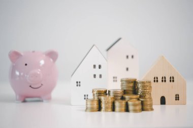 Ahşap ev modeli, bozuk para yığınları ve beyaz arka planda pembe kumbaralar, ev tasarruflarını, ipotek taksitlerini, emlak bütçesini ve finansal güvenlik hedeflerini temsil ediyor..