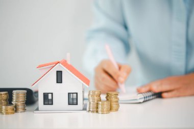 Masasında yığılı madeni paralar olan manken evi, ev bütçesi planlaması, ipotek ödemeleri, peşinat birikimleri ve emlak hedefleri ile fotokopi alanı hedeflerini gösteren kişi deftere yazarken.