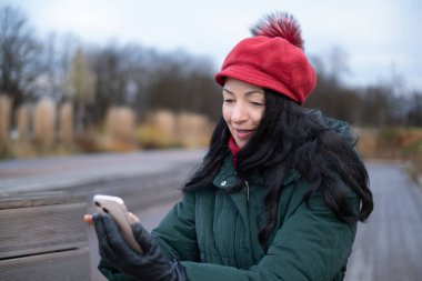 Kırmızı şapkalı ve yeşil ceketli yaşlı kadın açık havada akıllı telefonunu mutlulukla kullanıyor. Temiz havanın ve neşeyle gülümserken çevrenin tadını çıkarıyor.
