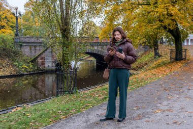 Kahverengi ceketli orta yaşlı bir kadın, elinde kahve fincanı olan sakin bir kanalın yanında duruyor. Etrafı canlı sonbahar yaprakları ve fotokopi alanı olan ağaçlarla çevrili.