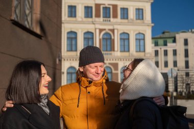 Güneşli bir günde, iki kadın ve bir erkek birlikte gülüyorlar, büyüleyici bir mimari zemin ile neşeli atmosferi güçlendiriyorlar.