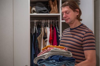 Erkek kişi, iyi organize edilmiş bir dolabın önünde ev işlerini ve ev idaresini ön plana çıkarırken bir yığın temiz çamaşır tutuyor.