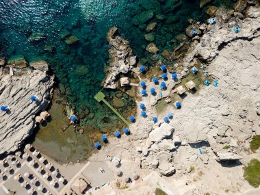 Kayalık bir sahil manzarası ve güneşlik ve şemsiyeli turkuaz bir deniz.