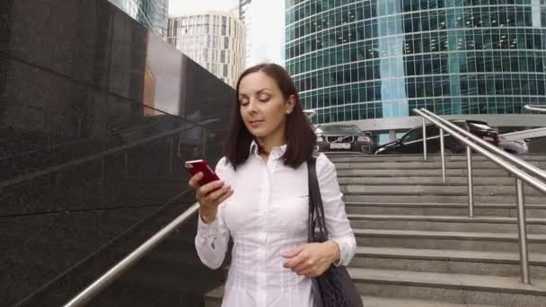 Menina bonita é mensagem de texto perto de escritórios de luxo e arranha-céus (steadicam tiro ) — Vídeo de Stock