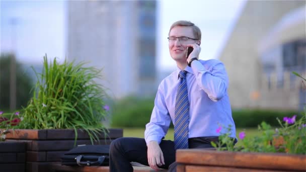 Χαρούμενος νεαρός επιχειρηματίας που μιλάει στο τηλέφωνο συνεδρίαση σε έναν πάγκο στο δρόμο — Αρχείο Βίντεο