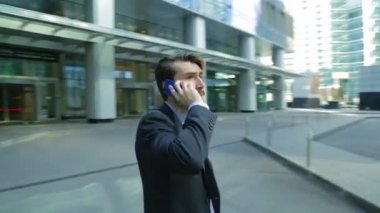 Yakışıklı işadamı modern iş merkezi yakınlarındaki akıllı telefonda konuşuyor gökdelen. Kamera adam (steadicam shot hareket ediyor)