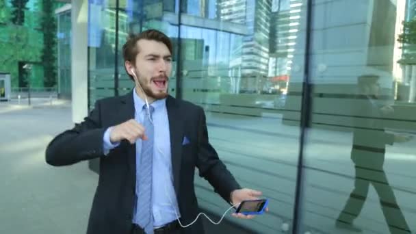 Pohledný muž nosí oblek poslech hudby do sluchátek, chytrý telefon, chůze, tanec, zpěv (steadicam shot) — Stock video