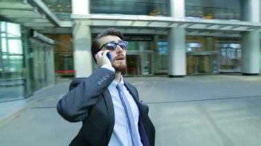 Güneş gözlüğü yakışıklı işadamı modern iş merkezi yakınlarındaki telefonda konuşuyor gökdelen. Kamera adam (steadicam shot hareket ediyor)