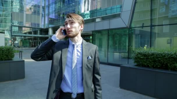 Hombre de negocios guapo está hablando en el teléfono inteligente cerca del moderno centro de negocios, rascacielos — Vídeo de stock