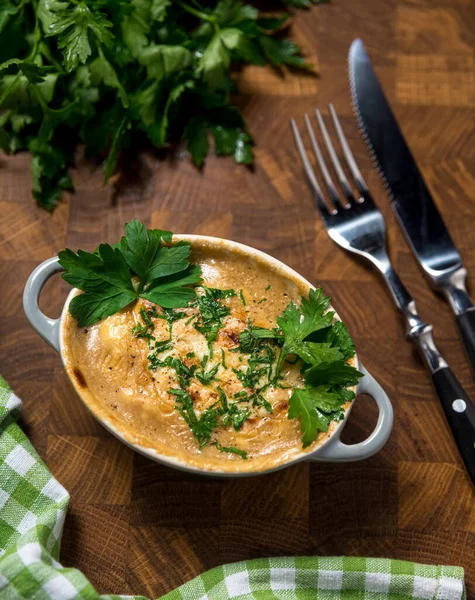 A small portion of creamy Russian julienne with mushrooms and cheese, garnished with parsley leaves. On a wooden table, a bunch of pepper, a knife and a fork. View from above. — Stock Image Small Portion Creamy Russian Julienne Mushrooms Cheese Garnished Parsley Leaves Royalty Free Stock Images