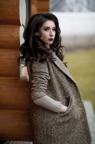 Primavera en el estilo country. Retrato de larga duración de mujer joven en suéter de punto blanco y sombrero peludo de pie en la parte delantera de la pared de madera rústica en el patio cerca de la casa, con suéter casual . — Foto de Stock