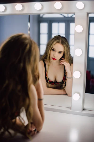 Молодая красивая женщина, нанося макияж лицо кистью, глядя в зеркало, сидя на стуле в гримерке с винтажным зеркалом темной комнате, носить красное белье — стоковое фото