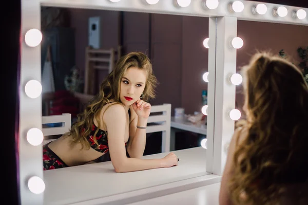 Joven hermosa mujer aplicando su maquillaje cara con cepillo, mirando en un espejo, sentado en la silla en el vestidor con espejo vintage habitación oscura, usando lencería roja — Foto de Stock