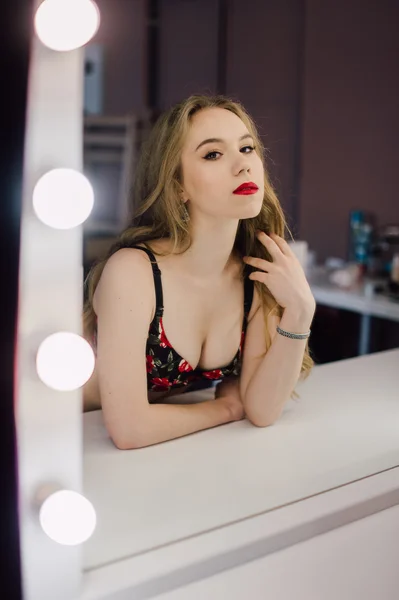 Jovem mulher bonita aplicando sua maquiagem rosto com escova, olhando em um espelho, sentado na cadeira no camarim com espelho vintage quarto escuro, vestindo lingerie vermelha — Fotografia de Stock