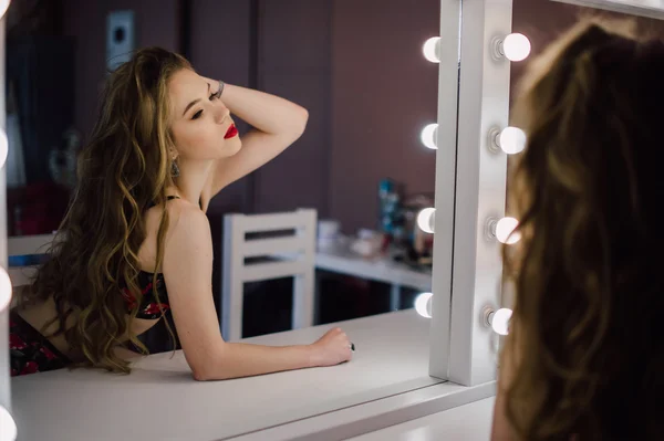 Mooie jongedame haar make-up gezicht met borstel toe te passen in een spiegel kijken, zittend op een stoel in de kleedkamer met vintage spiegel donkere kamer, het dragen van rode lingerie — Stockfoto