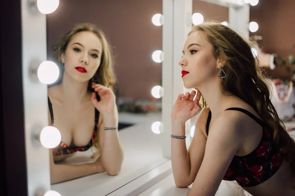Молодая красивая женщина, нанося макияж лицо кистью, глядя в зеркало, сидя на стуле в гримерке с винтажным зеркалом темной комнате, носить красное белье — стоковое фото