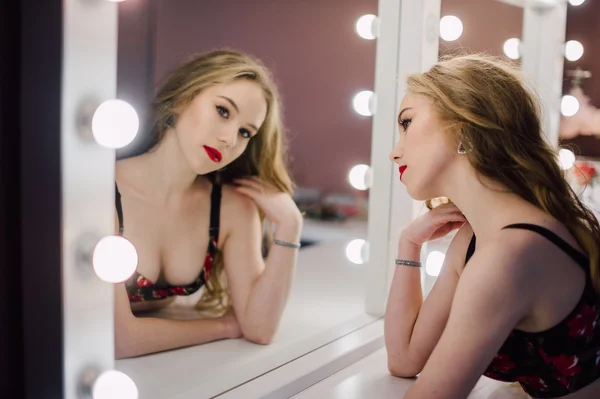 Mooie jongedame haar make-up gezicht met borstel toe te passen in een spiegel kijken, zittend op een stoel in de kleedkamer met vintage spiegel donkere kamer, het dragen van rode lingerie — Stockfoto