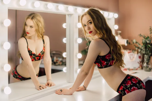 Νεαρή γυναίκα όμορφη εφαρμογή της make up προσώπου με βούρτσα, κοιτάνε στον καθρέφτη, κάθεται στην καρέκλα στο καμαρίνι με vintage καθρέφτη σκοτεινό δωμάτιο, φορώντας κόκκινα εσώρουχα — Φωτογραφία Αρχείου