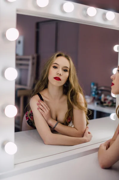 Молода красива жінка наносить макіяж обличчям пензлем, дивлячись у дзеркало, сидить на стільці в роздягальні з вінтажним дзеркалом темної кімнати, в червоній білизні — стокове фото