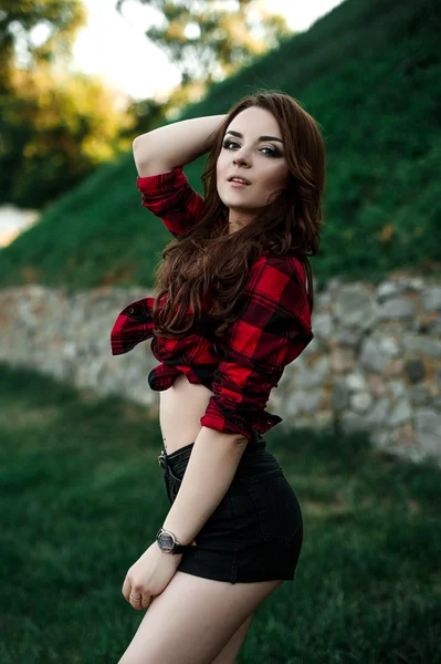 Hermosa chica hipster joven posando y sonriendo cerca de fondo de pared urbana en camisa a cuadros rojo, pantalones cortos, al aire libre retrato de verano . —  Fotos de Stock