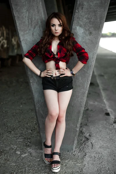 Hermosa chica hipster joven posando y sonriendo cerca de fondo de pared urbana en camisa a cuadros rojo, pantalones cortos, al aire libre retrato de verano . — Foto de Stock