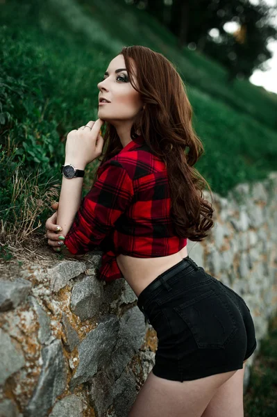 Hermosa chica hipster joven posando y sonriendo cerca de fondo de pared urbana en camisa a cuadros rojo, pantalones cortos, al aire libre retrato de verano . — Foto de Stock