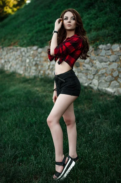 Hermosa chica hipster joven posando y sonriendo cerca de fondo de pared urbana en camisa a cuadros rojo, pantalones cortos, al aire libre retrato de verano . — Foto de Stock