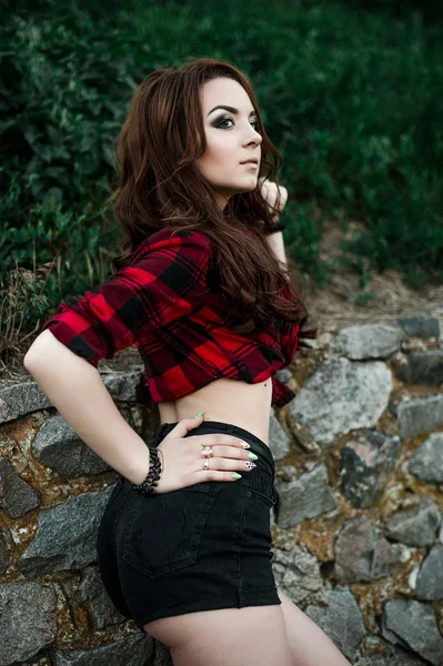 Hermosa chica hipster joven posando y sonriendo cerca de fondo de pared urbana en camisa a cuadros rojo, pantalones cortos, al aire libre retrato de verano . — Foto de Stock