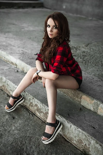 Hermosa chica hipster joven posando y sonriendo cerca de fondo de pared urbana en camisa a cuadros rojo, pantalones cortos, al aire libre retrato de verano . — Foto de Stock