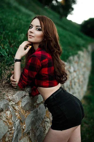 Bella giovane ragazza hipster posa e sorridente vicino al muro urbano sfondo in camicia a quadri rossa, pantaloncini, all'aperto ritratto estivo . — Foto Stock