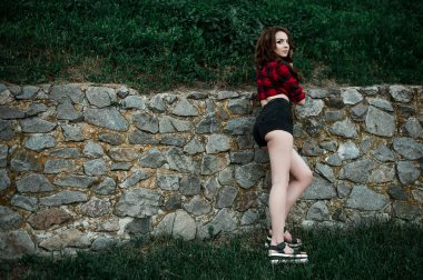 Poz ve kentsel duvar arka planda kırmızı kareli gömlek, şort, açık havada yaz portre yakınındaki gülümseyen güzel genç hippi kız.