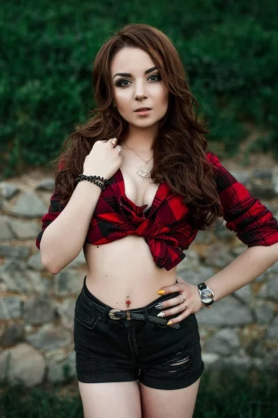 Poz ve kentsel duvar arka planda kırmızı kareli gömlek, şort, açık havada yaz portre yakınındaki gülümseyen güzel genç hippi kız.
