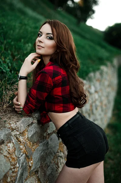 Beautiful young hipster girl posing and smiling near urban wall background in red plaid shirt, shorts, outdoors summer portrait. — Stock Photo, Image